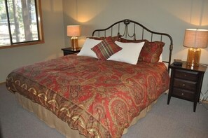 Uma cama com colcha estampada em vermelho e dourado, duas mesas de cabeceira com abajures, e uma janela com vista para árvores.