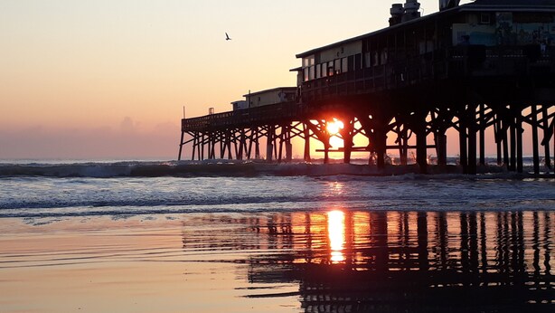 Eine Steg, der sich ins Meer erstreckt, mit untergehendem Sonnenuntergang im Hintergrund, der sich im feuchten Sand spiegelt.