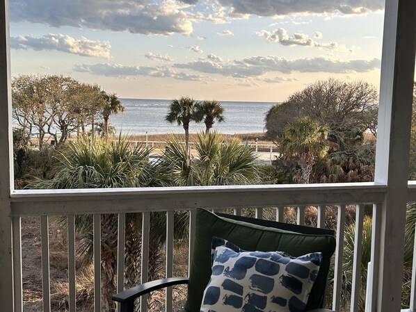 A view from a porch with a chair and a cushion, overlooking a beach with palm trees and a fence.