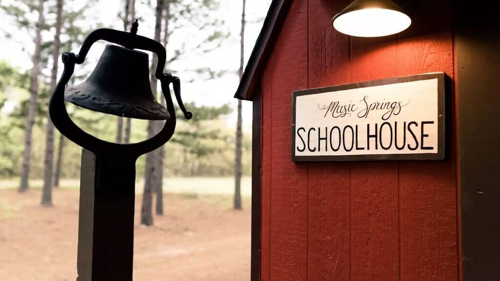Eine Glocke und ein Schild mit der Aufschrift 'Music Springs SCHOOLHOUSE' vor einem Holzhintergrund.