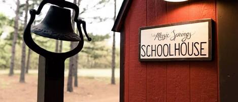 Eine Glocke und ein Schild mit der Aufschrift 'Music Springs SCHOOLHOUSE' vor einem Holzhintergrund.