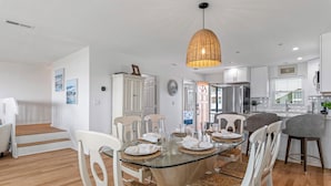 A modern kitchen with white cabinets, a stainless steel refrigerator, and a dining area with a glass table and wicker chairs. - Luxury Oceanfront Retreat: Concierge Beach Tent Service, Heated Pool & Hot Tub (Holden Beach)