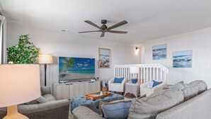 A modern living room with a grey sofa, a white armchair, a ceiling fan, and a view of the ocean through the window. - Luxury Oceanfront Retreat: Concierge Beach Tent Service, Heated Pool & Hot Tub (Holden Beach)