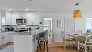 A modern kitchen with white cabinets, a central island, and a dining area with white chairs and a glass table. - Luxury Oceanfront Retreat: Concierge Beach Tent Service, Heated Pool & Hot Tub (Holden Beach)