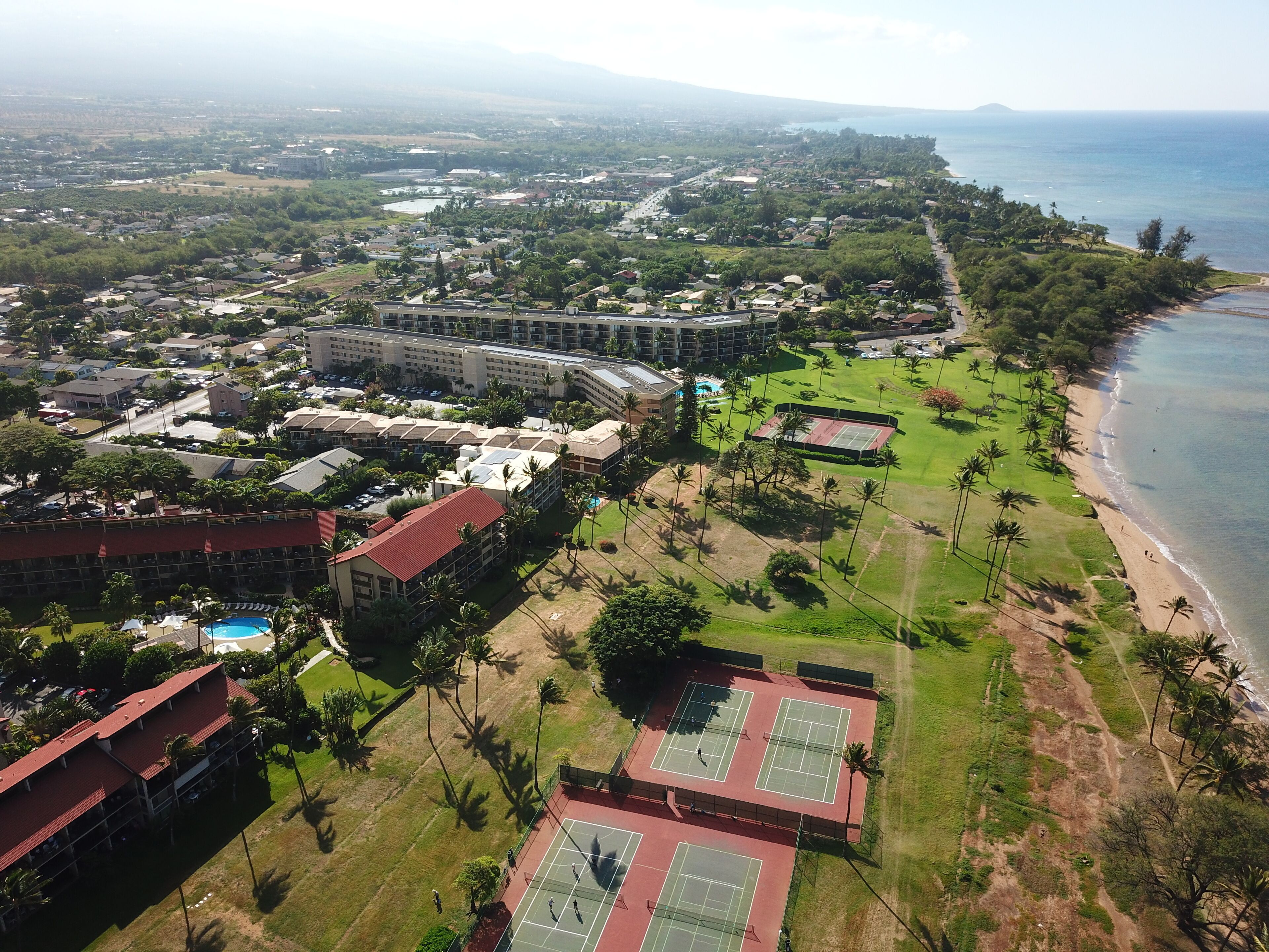 An aerial view of a coastal resort with multiple buildings, a beach, and tennis courts.