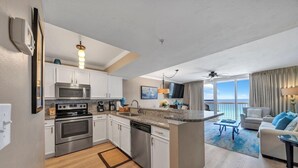 Una cocina moderna con electrodomésticos de acero inoxidable, un comedor con alfombra azul y una sala de estar con vista al océano.