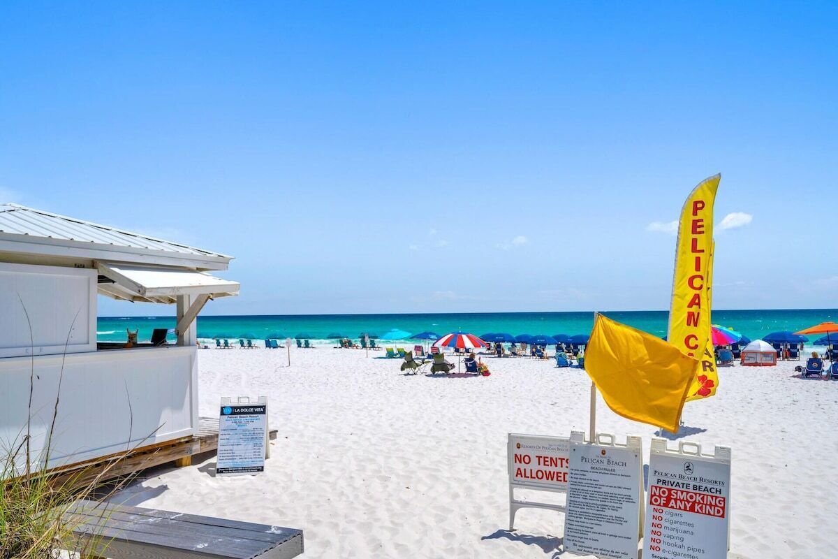 A beach scene with a no tent allowed sign, a no smoking sign, and a yellow flag with the word "PELICAN" on a beach umbrella.