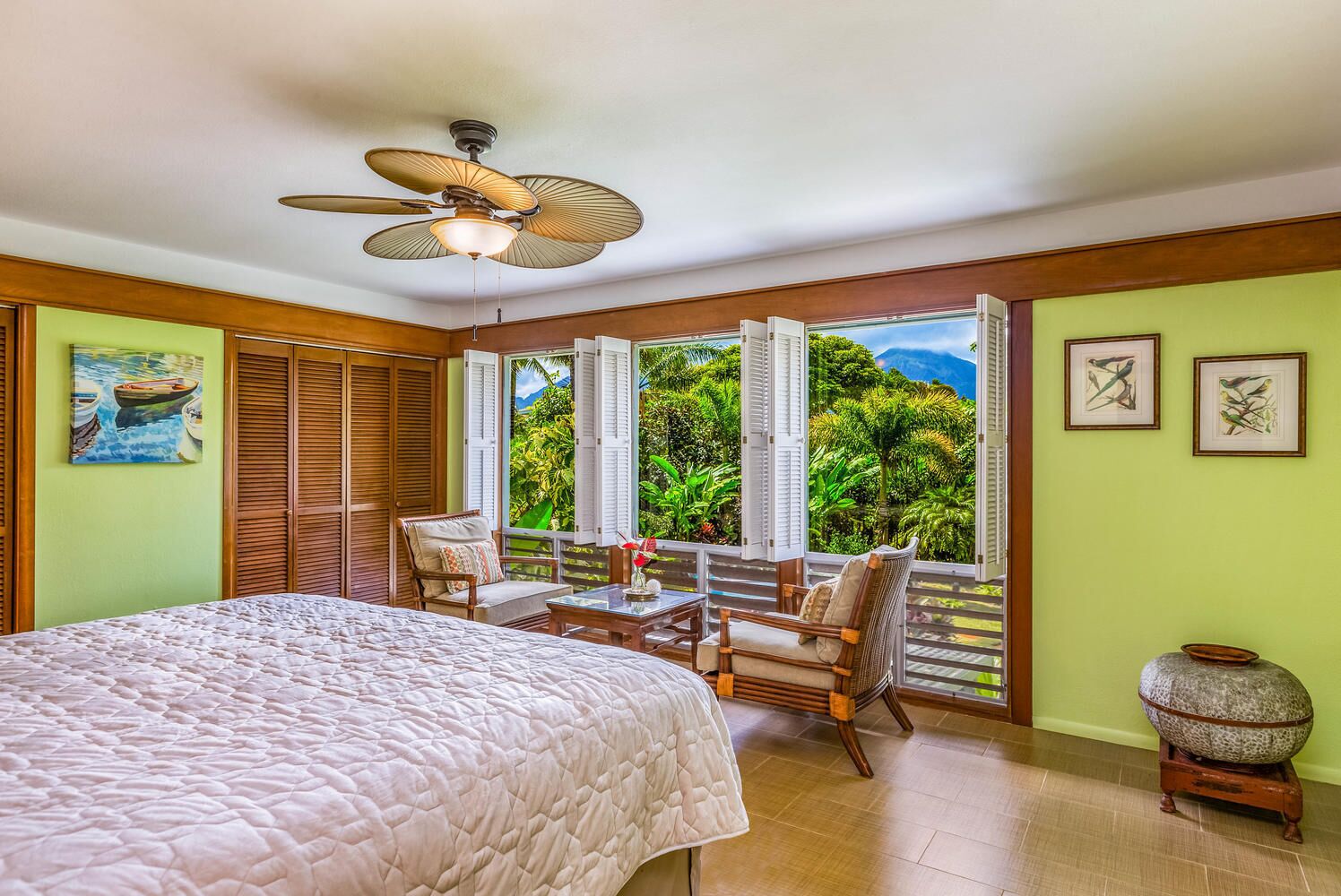 A bedroom with a bed, a ceiling fan, a window with shutters, a table with chairs, and a view of greenery outside.