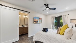 A bedroom with a bed, a ceiling fan, a window with curtains, a bathroom with a mirror, and a painting on the wall. - Walk to Old Town, 3BR + Backyard (Scottsdale)