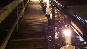 Eine Holzpromenade führt zu einem Gebäude mit mehreren Fenstern und einem Balkon, umgeben von Wasser und Palmen.