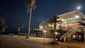 Ein zweistöckiges Strandhaus mit Balkon, Palmen und einer Treppe, die zum Obergeschoss führt.