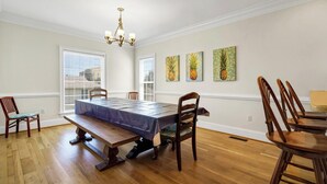 Une salle à manger avec une longue table, des chaises en bois et un lustre. Trois tableaux ornent le mur, et les fenêtres sont équipées de stores.