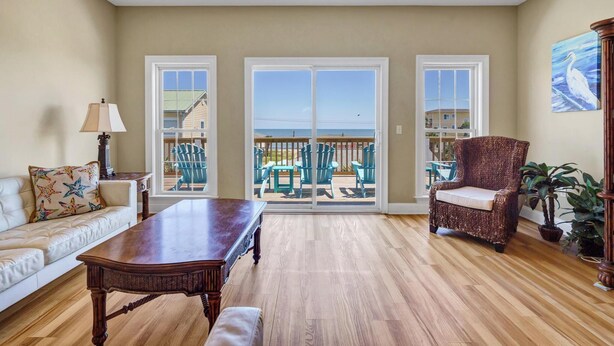 Un salon spacieux doté d’un plancher en bois, d’un canapé, d’une table et d’une chaise. De grandes fenêtres offrent une vue sur la plage, et un balcon est aménagé avec du mobilier d’extérieur.
