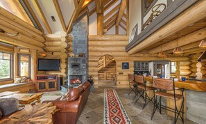 A log cabin interior with a stone fireplace, wooden furniture, and a bar area.