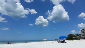 Une plage de sable blanc, un ciel bleu azur et quelques nuages. On y trouve des parasols et des chaises longues.