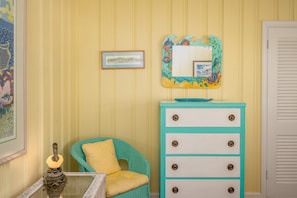 Una habitación con un aparador azul verdoso, una silla de mimbre con cojín amarillo y un cuadro en la pared.