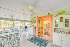A room with a ceiling fan, a dining area with tables and chairs, a wicker chair, a cabinet with decorative items, and a door leading to another room. - Spacious Beach Home for the Whole Fam, Steps from the Sand by Tybee Cottages (Tybee Island)