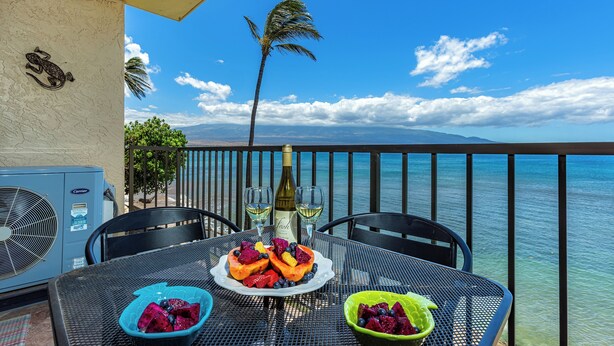 Une table à manger avec une salade de fruits tropicaux, une bouteille de vin et un bol de myrtilles, le tout sur un balcon donnant sur l’océan.