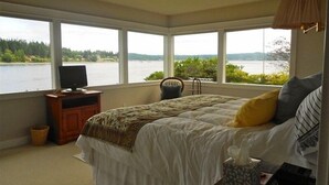 A bedroom with a large bed, a television on a wooden cabinet, and a view of a lake through large windows. - Bainbridge Island (Puget Sound):No-Bank Waterfront Sandy Beach (Bainbridge Island)