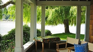 A covered outdoor seating area with a table and chairs, overlooking a lake with trees. - Bainbridge Island (Puget Sound):No-Bank Waterfront Sandy Beach (Bainbridge Island)