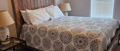 A bedroom with a wooden headboard, a patterned bedspread, two bedside lamps, and a window with blinds.