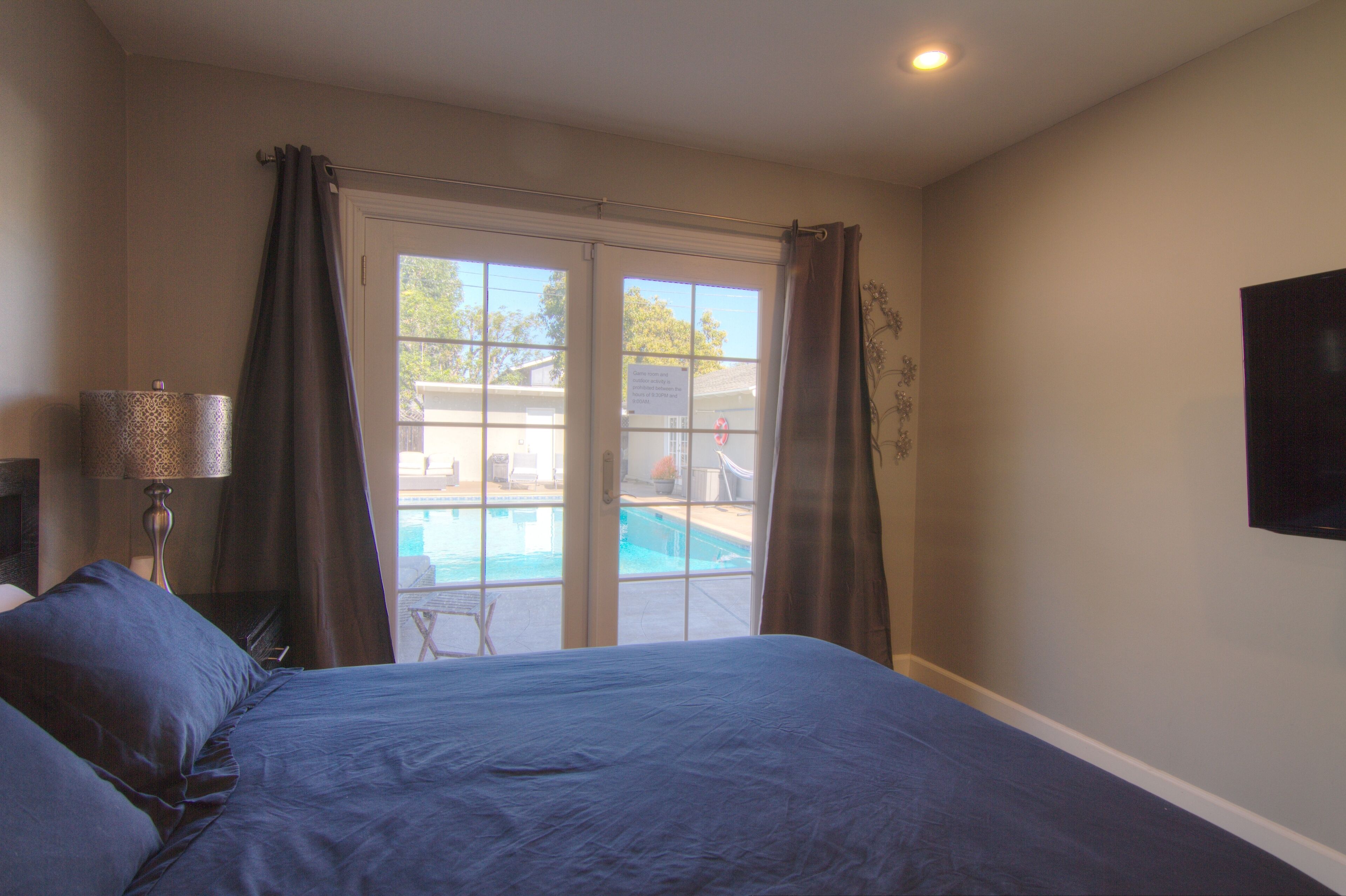 A bedroom with a large bed, a TV mounted on the wall, and a view of an outdoor swimming pool through a sliding glass door.