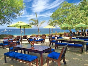 Een café aan het strand met houten tafels en blauw gestoffeerde stoelen, omringd door palmbomen en parasols, met uitzicht op de oceaan.