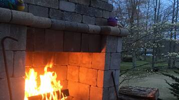 A stone fireplace with a fire burning inside, surrounded by logs and a bench.