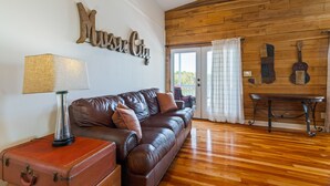 Un salon avec un canapé en cuir marron, une lampe, une valise et une œuvre d’art murale représentant une guitare.