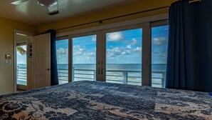 Une chambre dotée d’un grand lit, d’un ventilateur de plafond et offrant une vue sur l’océan grâce à de grandes portes-fenêtres coulissantes.