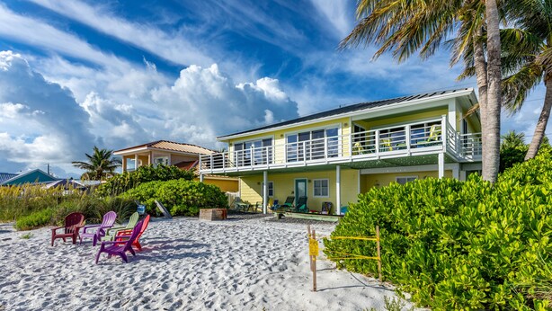 A beachfront property with a two-story house, a sandy beach, and several Adirondack chairs. - LABOR DAY SPECIALS ,BEACHFRONT, PRIVATE BEACH ON THE WATERFRONT, Bring your Dog (Anna Maria)
