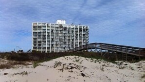 Um edifício moderno com telhado plano e várias varandas, ao lado de uma praia arenosa com uma passarela de madeira.