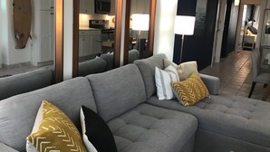 Uma sala de estar moderna com um sofá modular cinza, uma decoração com prancha de surf e uma cozinha ao fundo.