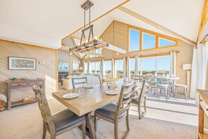 En rummelig spiseplads med et træbord og stole, store vinduer der giver udsigt over landskabet, og et højt loft med synlige bjælker.