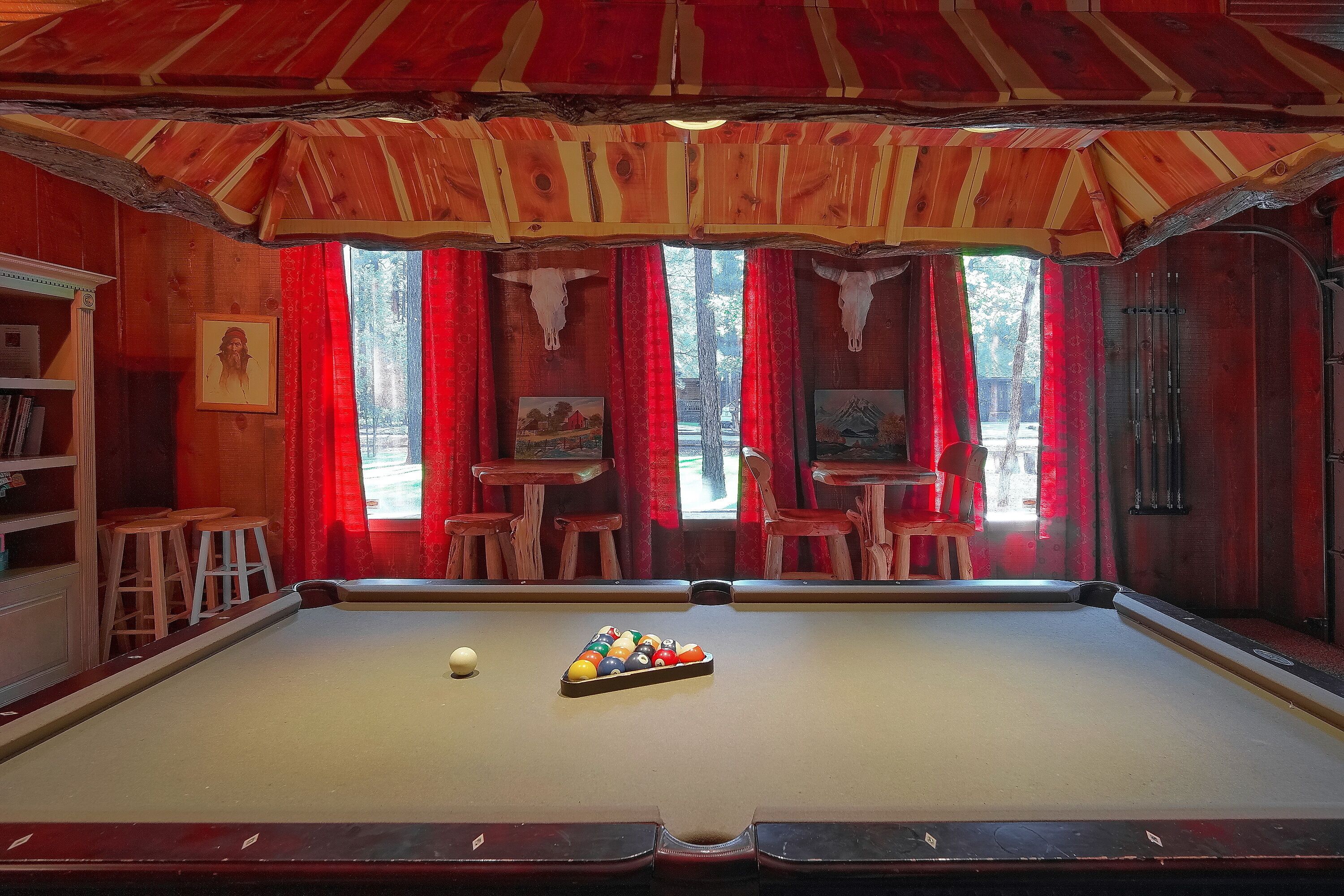 A pool table with a cue ball and a rack of billiard balls, wooden stools, a bookshelf, and red curtains.