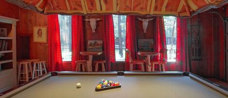 A pool table with a cue ball and a rack of billiard balls, wooden stools, a bookshelf, and red curtains.