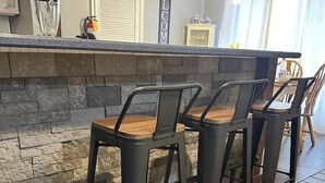 A kitchen with a stone backsplash, two metal bar stools, a wooden countertop, and a 'WELCOME' sign.