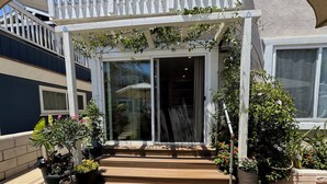 Een overdekte ingang met een houten terras dat leidt naar een glazen deur, omringd door potplanten en een klein vloerkleed.
