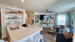 Een moderne keuken met een centraal eiland, een eethoek met een ronde tafel, een woonkamer met een bank en een televisie, en een plafondventilator.