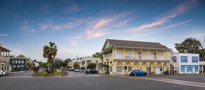 A street view of a town with a yellow building with a balcony, parked cars, and palm trees. - Hang up all your cares and woes and head to Southern Bell with Riverfront views! (St Marys)