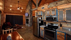 A rustic cabin interior with a stone fireplace, wooden ceiling beams, a dining area with a table and chairs, and a kitchen with modern appliances and blue-framed cabinets. - Ole Blue (Broken Bow)