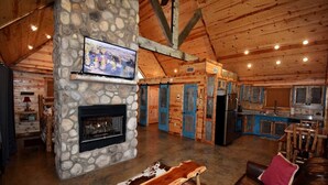 A cozy cabin interior with a stone fireplace, a television mounted on a stone wall, a kitchen area, and a seating area with a leather couch and a wooden bench. - Ole Blue (Broken Bow)