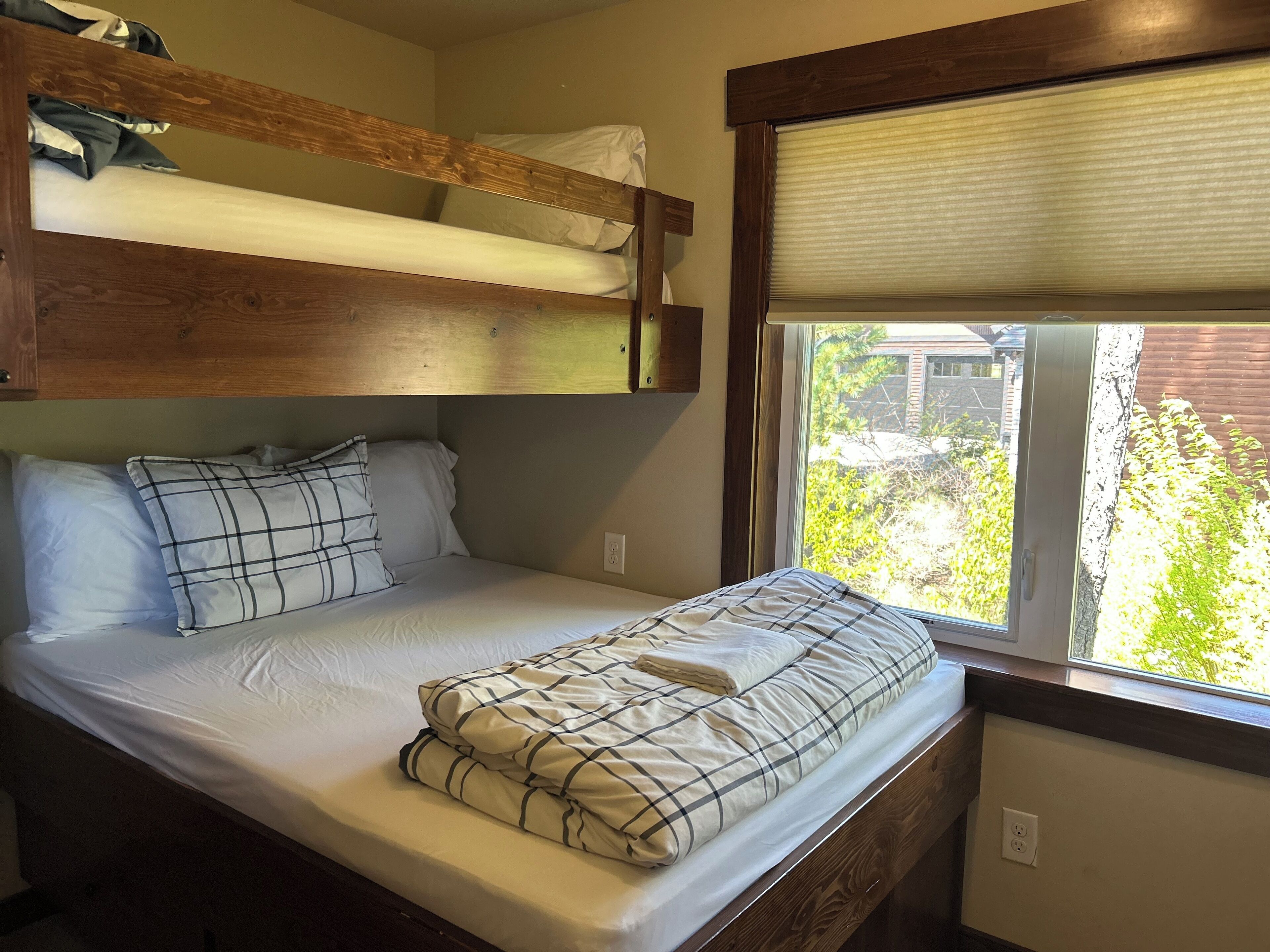 A bunk bed with a window above the lower bunk, a bedside table, and a view of greenery outside.