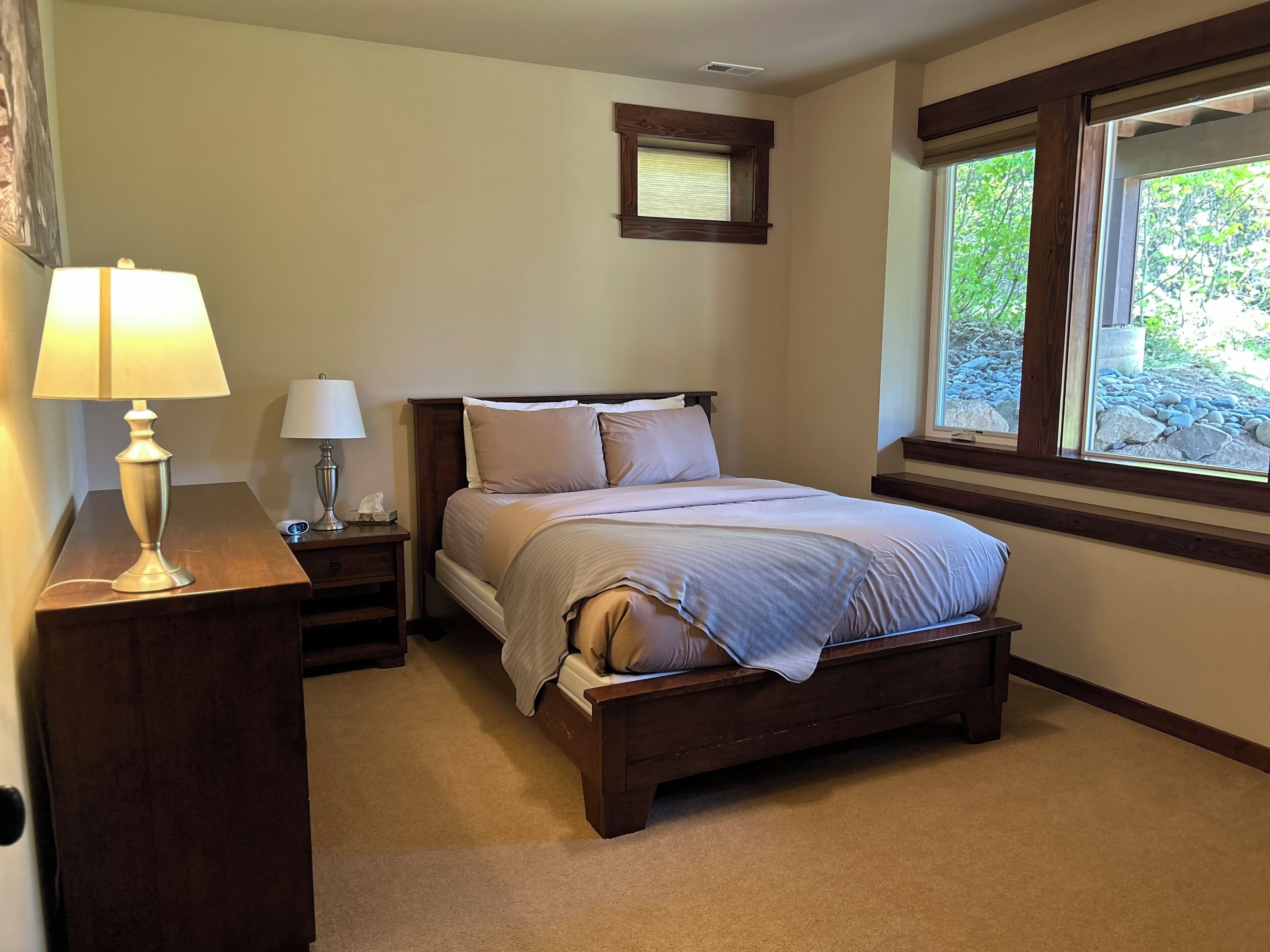 Une chambre à coucher avec un lit en bois, deux tables de chevet avec des lampes, une fenêtre donnant sur un paysage verdoyant et une petite fenêtre située au-dessus du lit.
