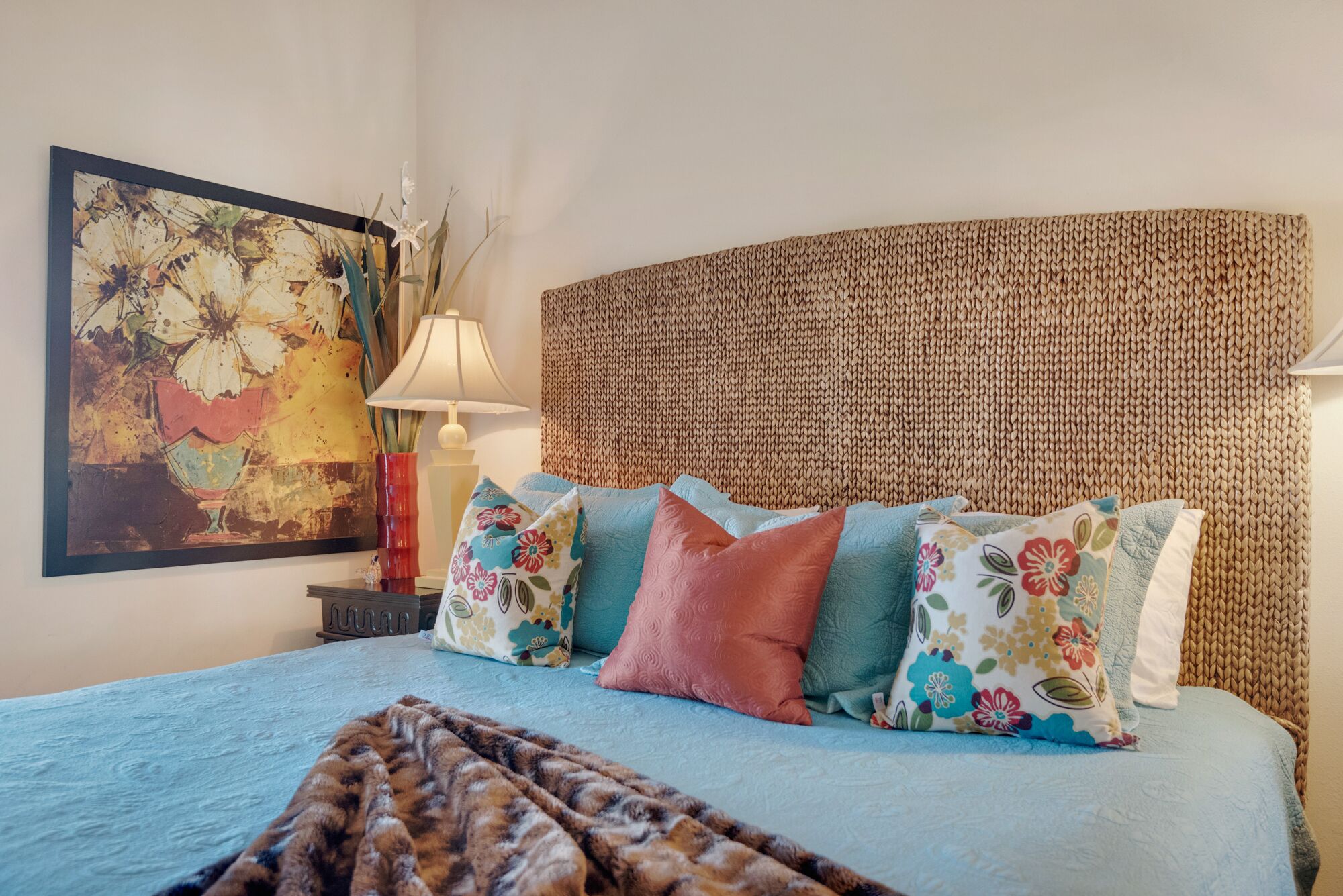 Een slaapkamer met een bekled hoofdbord, een bed met bloemenkussens, een decoratieve lamp en een schilderij aan de muur.