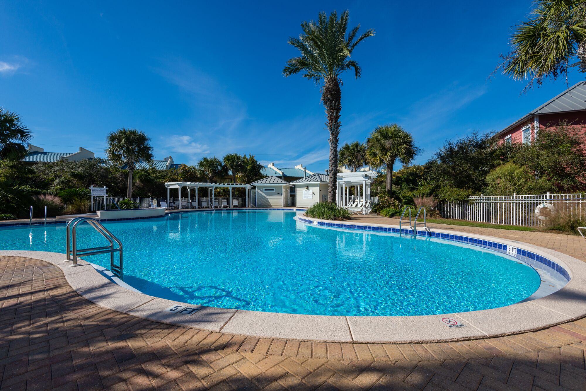 A clear blue swimming pool with a palm tree in the center, surrounded by lounge chairs and a covered seating area.