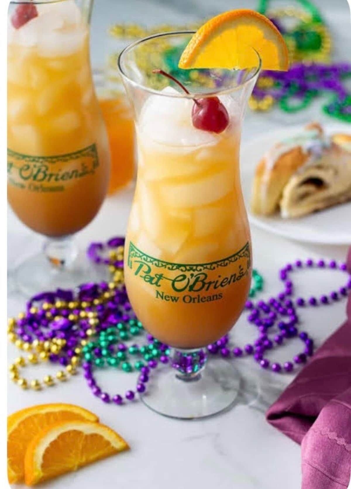 Una copa de cóctel con una cereza encima, rodeada de collares de Mardi Gras y una rodaja de naranja.