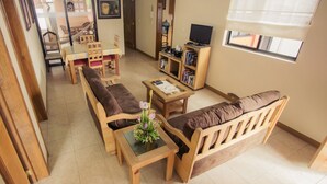 A cozy living room with a sofa, a coffee table, and a TV. There's a bookshelf and a dining area with chairs and a table.