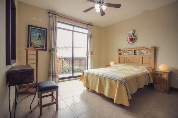 A bedroom with a bed, a desk, a chair, a ceiling fan, and a balcony with a view.