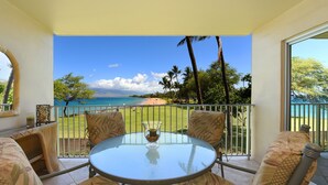 A balcony with a glass table and four chairs, overlooking a beach with palm trees and a clear blue sky. - Beautiful 2 Bdrm Oceanfront Kihei Condo!  (Kihei)